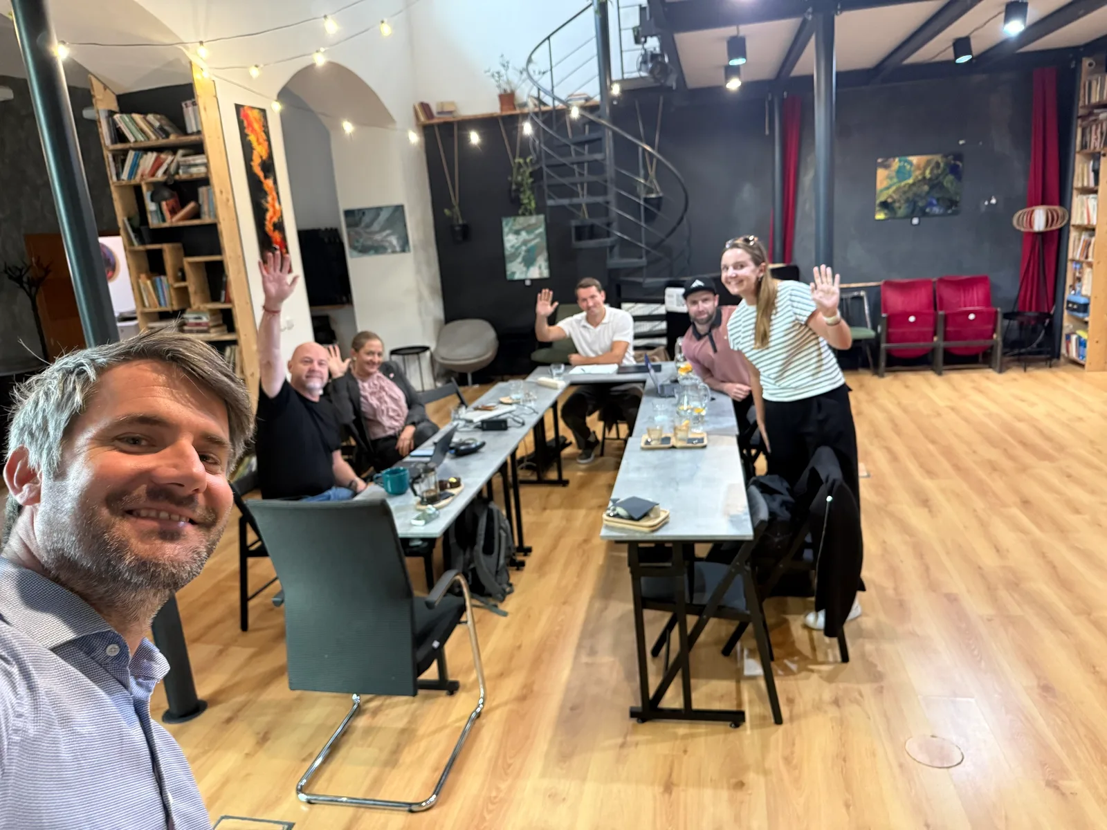 Účastníci workshopu Stavíme s AI u stolů v Coworkingu Pracovna na Žižkově, mávají do fotoaparátu